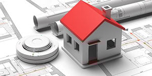 red roofed toy house on building plans with a fire detector safety alarm
