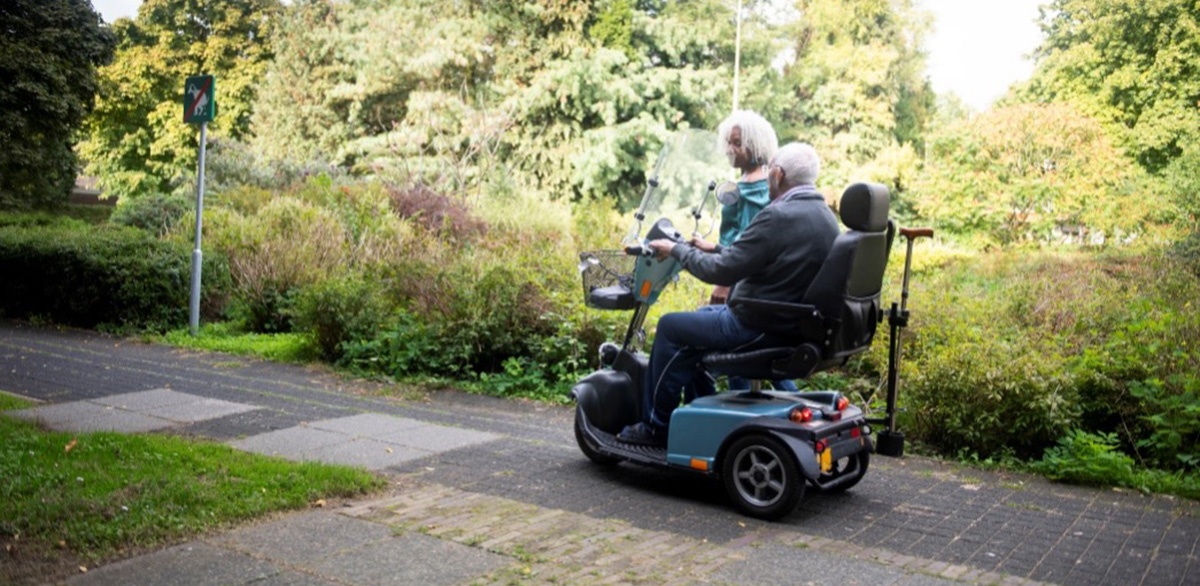 A woman walking alongside a man on a mobility scooter.