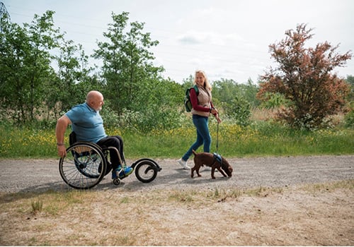 A man in a wheelchair with a woman walking a dog outdoors | Mobility Blog